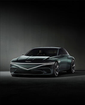 The Genesis X Speedium Coupe Concept vehicle, featuring a dark greenish tint, is parked in a darkened interior. It is bathed in white light, with the front lights turned on. The top row of two front lights extends horizontally, while the bottom row forms a gentle V-shape at the center of the car’s front.